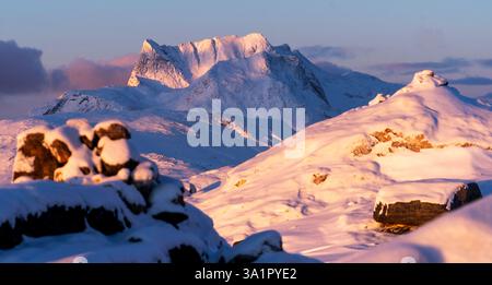 Un paysage hivernal à couper le souffle avec des montagnes enneigées sous une douce lueur de coucher de soleil. Le premier plan comprend des formations rocheuses partiellement couvertes Banque D'Images