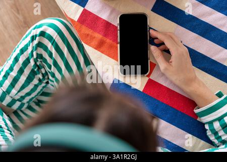 Vue de dessus d'une personne portant un pyjama rayé vert, tenant un smartphone avec écran noir sur une couverture rayée colorée, scène de maison confortable et décontractée Banque D'Images