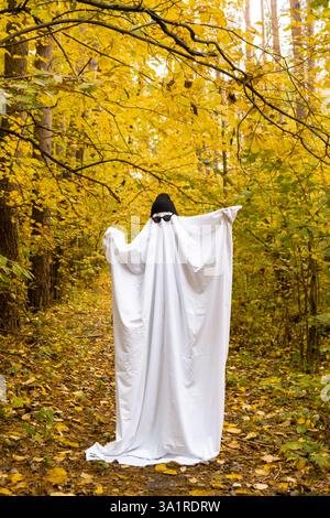 Joyeuses vacances d'Halloween. Mignonne personne méconnaissable dansant en costume de fantôme sur fond de forêt d'automne. Costume créatif avec chapeau blanc et noir et lunettes. Concept minimal Banque D'Images