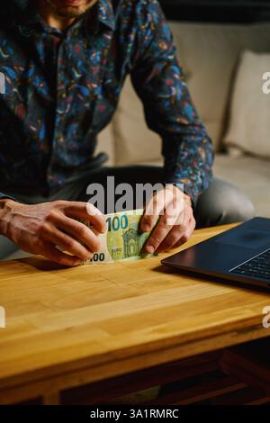Homme tenant cent billets de banque en euros, assis sur un canapé devant la table avec un ordinateur portable ouvert. Personne comptant l'argent comptant à la maison. Finances personnelles et h Banque D'Images