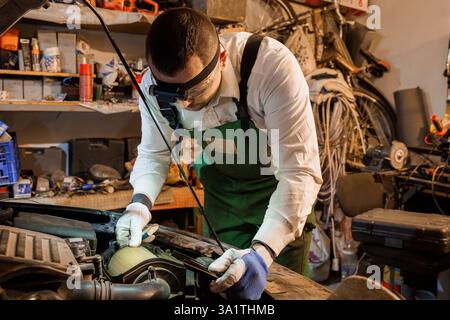 Mécanicien concentré portant phare et gants réparant le moteur de voiture dans un environnement d'atelier encombré. Banque D'Images