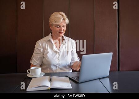 Belle dame aux cheveux blonds habillée en chemise blanche tout en étant assise à table et travaillant sur un ordinateur portable, femme professionnelle engagée dans une activité ciblée Banque D'Images