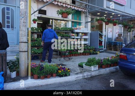 Un homme tient un tuyau et arrose les fleurs et les plantes dans des pots qu'il a placés sur le trottoir à l'extérieur de son magasin Banque D'Images