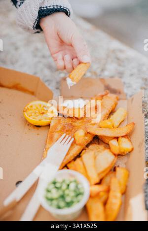 Une personne mange du poisson et des frites dans une boîte en carton. Le poisson est recouvert de pâte et les chips sont dorés. Il y a une fourchette et un couteau sur le Banque D'Images