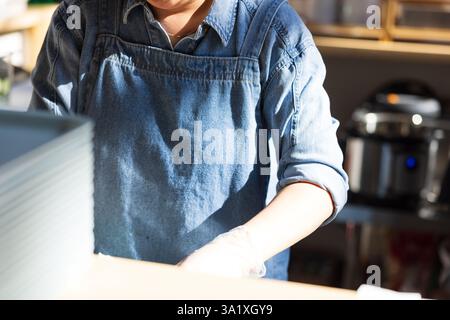 Une femme dans un tablier bleu travaille dans une cuisine. Elle porte des gants et prépare de la nourriture Banque D'Images