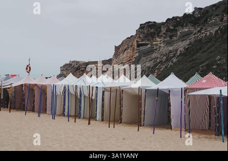 Des tentes de plage rayées colorées se dressent sur la rive sablonneuse de Praia da Nazaré Banque D'Images