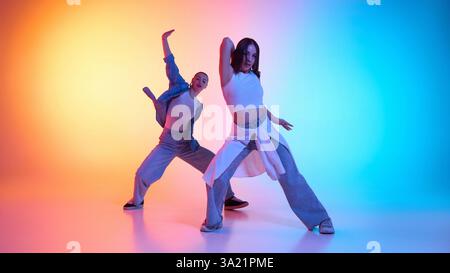 Deux jeunes femmes énergiques dans des tenues décontractées, dansant hip hop sous néon gradient lueur en arrière-plan studio. Banque D'Images