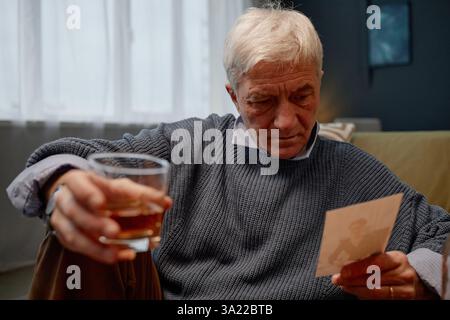Homme senior assis sur le canapé, tenant un verre et regardant une vieille photographie, enveloppé dans une expression réfléchie, portant un pull, la lumière du soleil filtrant à travers la fenêtre en arrière-plan Banque D'Images