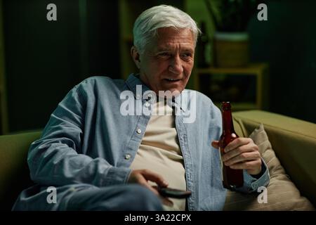Homme âgé assis sur le canapé tenant la télécommande et la bouteille de bière tout en regardant quelque chose à la télévision, profitant d'un moment de détente à la maison, portant une chemise décontractée avec une expression neutre Banque D'Images