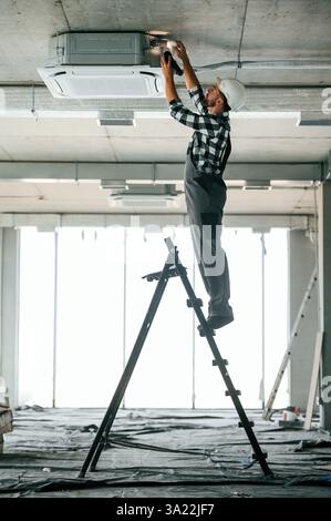Debout sur l'échelle. Réparateur en uniforme est dans le bâtiment inachevé. Banque D'Images