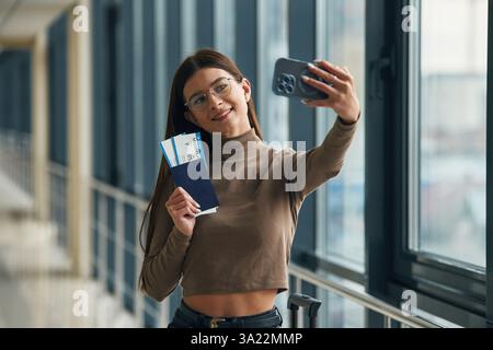 Faire selfie avec le téléphone, sourire. Jeune femme voyageuse dans le hall d'entrée de l'aéroport. Banque D'Images