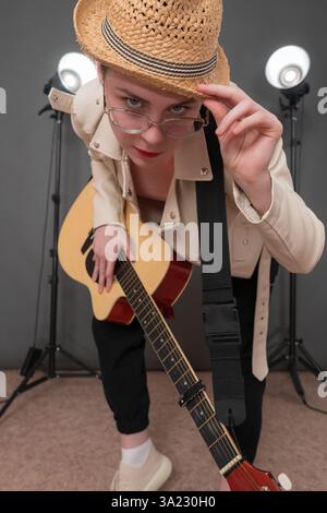 Guitariste féminine dans les lunettes tient la guitare et regarde avec confiance la caméra, s'inclinant devant la caméra et se pliant magnifiquement tout en tenant la guitare. D'une main Banque D'Images