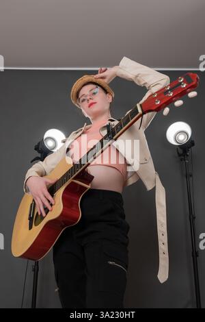 Portrait de guitariste féminine confiante tenant la guitare, regardant la caméra. D'une main, elle tient le bord de son chapeau de paille, sa tenue de scène complétée Banque D'Images