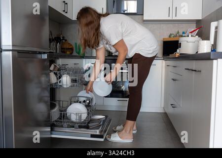 Femme déchargeant la vaisselle propre d'un lave-vaisselle ouvert dans une cuisine moderne avec des armoires blanches et des appareils en acier inoxydable. Concept des tâches ménagères Banque D'Images
