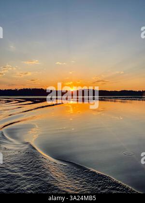 Un coucher de soleil doré se reflète sur la surface calme du lac, avec de douces ondulations façonnant l'eau. Le ciel passe de l'orange au bleu, créant un cadre paisible Banque D'Images