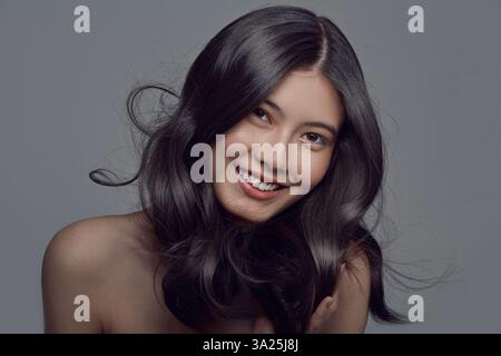 Jolie femme asiatique aux cheveux longs et brillants ondulés sur un gris isolé. Banque D'Images