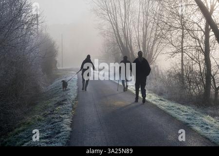 Sur le chemin de Radley Boathouse par la Tamise. Un de mes sentiers préférés, n'importe où. Il mène de mon village natal de Radley dans l'Oxfordshire, en Angleterre, à une partie pittoresque de la Tamise près de Radley Boathouse. Il parvient à paraître joli tout au long de l'année - et en particulier ici un matin d'hiver brumeux, juste après le lever du soleil. Un promeneur de chien et des joggers disparaissent dans les brumes... Banque D'Images