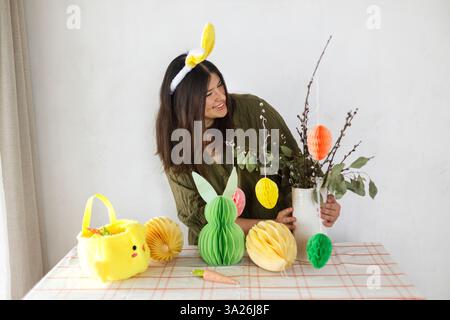 Joyeuses Pâques ! Femme élégante dans les oreilles de lapin décorant avec des œufs de papier de pâques colorés, lapin et carottes sur la table. Table de Pâques et chasse à Pâques Banque D'Images