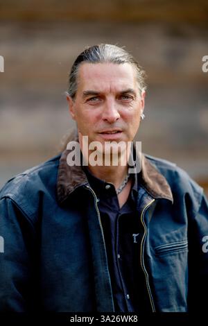 Homme aux cheveux longs dans une veste sombre se tient à l'extérieur avec un fond flou. Brandebourg, Allemagne Banque D'Images