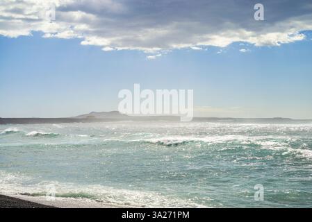 Vagues océaniques s'écrasant sur une rive rocheuse sous un ciel nuageux avec des montagnes lointaines. Lanzarote, Espagne Banque D'Images