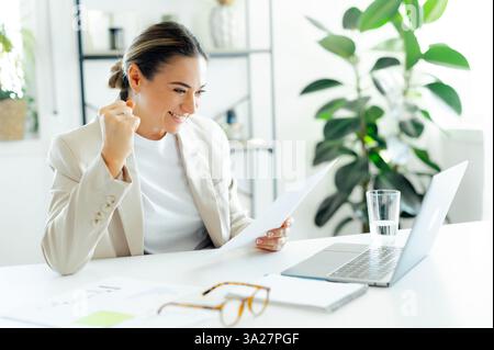 Joyeuse femme d'affaires caucasienne réussie dans un blazer beige, célèbre le succès tout en lisant un document à son bureau avec un ordinateur portable, l'air joyeuse, a obtenu de bons résultats de travail, sourit Banque D'Images