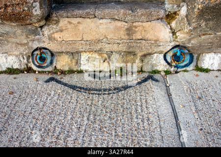 Un visage souriant dessiné sur un chemin en béton, avec des yeux positionnés autour de vieux anneaux d'amarrage rouillés Banque D'Images