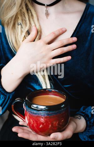 Femme blonde avec des dreadlocks tenant la tasse de cacao et la main sur le coeur, portant le collier pendentif serpent Banque D'Images