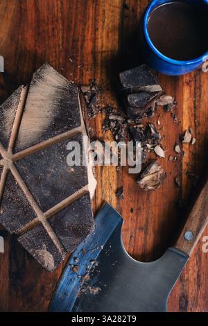 Préparation cérémonielle de cacao avec bloc de cacao cru et cacao chaud dans une tasse en céramique sur planche de bois foncé avec hache de cuisine Banque D'Images