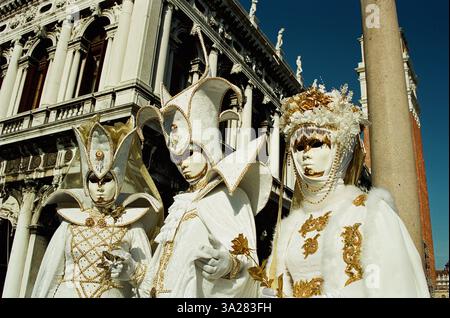 Trois personnages élégamment habillés dans des costumes et des masques élaborés, debout devant un bâtiment historique sous un ciel bleu clair. Les costumes sont Ado Banque D'Images