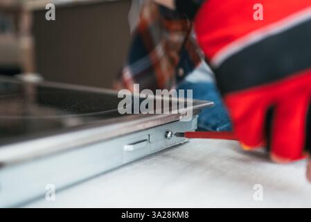 Main gantée de réparateur vissant la vis sur une plaque vitrocéramique lors de travaux d'entretien dans une cuisine, avec un propriétaire flou en arrière-plan Banque D'Images