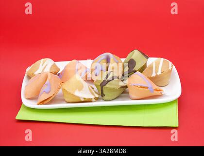 Gros plan sur une assiette de biscuits de fortune chinois arrosés de bonbons au chocolat colorés fondus, couchés sur une serviette verte sur fond rouge. Banque D'Images