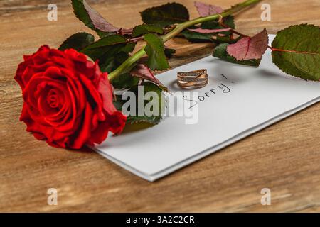 Une seule rose rouge repose sur une table en bois à côté d'une enveloppe blanche qui indique désolé, avec deux anneaux de mariage placés sur le dessus, symbolisant un engagement cassé Banque D'Images