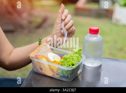 Vue latérale gros plan femme d'affaires mangeant de la salade de sa boîte à lunch assise sur le banc au parc de bureaux. Banque D'Images