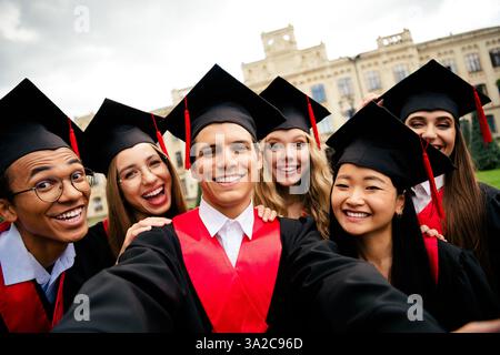 Groupe joyeux de jeunes diplômés célébrant le succès dans le cadre du campus extérieur avec mortarboards et diplômes sous un ciel d'été lumineux Banque D'Images