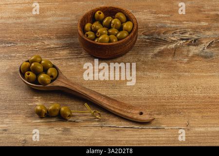 Les olives vertes sont disposées sur une surface en bois avec une cuillère en bois et un bol. Le cadre rustique met en valeur les textures et les couleurs naturelles de t Banque D'Images