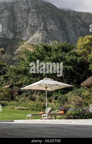 un parasol blanc jette une ombre sur deux chaises longues rembourrées. En arrière-plan, un jardin luxuriant et verdoyant s'étend et la montagne au loin, Tranqu Banque D'Images