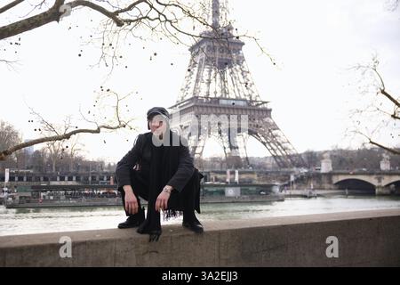 Un homme à la mode dans des vêtements sombres squats sur un rebord de pierre avec la Tour Eiffel derrière lui. Des branches d'arbres sans feuilles encadrent la scène hivernale parisienne. Banque D'Images