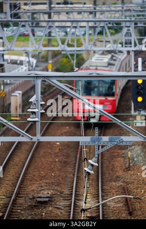 Le train de banlieue rouge à franges blanches passe sous des structures de réseau en aluminium pour les lignes de train pendant les heures de pointe, sur des voies argentées sur un lit de roche brun foncé. Banque D'Images