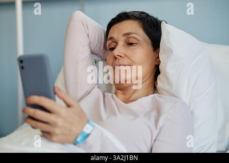 Femme âgée allongée dans le lit, tenant un smartphone avec la main droite et reposant sa tête sur des oreillers, paraissant confortable et détendue à l'hôpital ou en milieu de soins à domicile Banque D'Images
