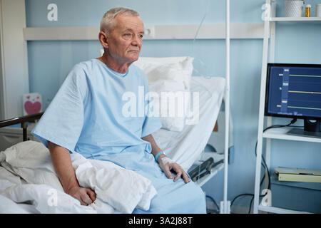 Homme senior reposant dans un lit d'hôpital, entouré d'équipements et de dispositifs médicaux, dans une pièce de couleur claire, regardant contemplative, soulignant l'environnement de soins de santé Banque D'Images