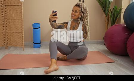 Femme prenant un selfie à la salle de gym avec des dreadlocks, assise sur un tapis à côté de balles d'exercice, mettant en valeur des tatouages et portant des vêtements de sport, souriant et c Banque D'Images