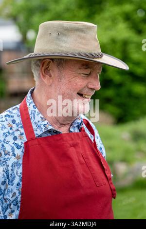Portrait d'un homme âgé souriant dans un chapeau de cow-boy et un tablier rouge lors d'un événement en plein air. Ambiance décontractée et joyeuse à la campagne, concept de retraite, c Banque D'Images