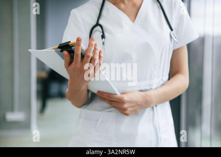 Debout et tenant des documents dans les mains. Médecin féminin en manteau blanc est à l'intérieur. Banque D'Images