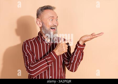 Portrait d'un homme âgé avec une chemise rayée élégante, souriant et faisant des gestes heureux devant un fond beige pointant vers sa paume. Banque D'Images
