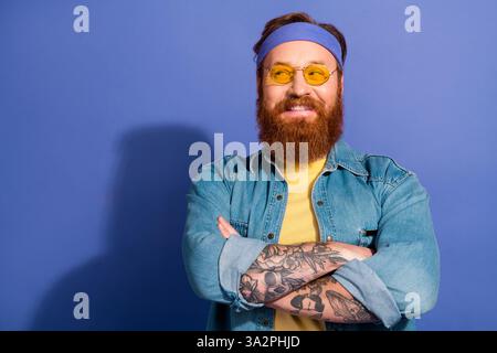 Homme barbu confiant dans une tenue décontractée en denim souriant sur fond violet, mettant en valeur le style rétro et hipster avec des tatouages. Banque D'Images