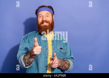 Homme adulte barbu souriant avec des tatouages pointant les mains joyeusement portant chemise décontractée en denim, bandeau, devant un fond violet Banque D'Images