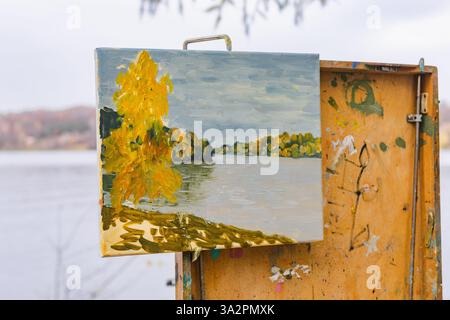 Une peinture de paysage d'automne représentant un arbre doré et une rivière calme exposée sur un chevalet en bois à l'extérieur Banque D'Images