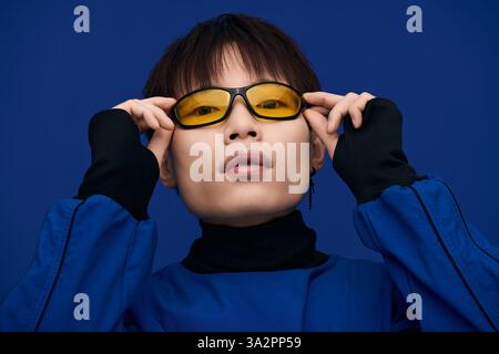 Un beau jeune homme asiatique respire le charme dans des lunettes de soleil jaunes frappantes sur un fond bleu frais. Banque D'Images