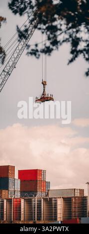 Un vertorama avec une grande grue industrielle soulevant un épandeur robuste au-dessus d'une cour de conteneurs d'expédition. Conteneurs rouges et bleus empilés, fil barbelé f Banque D'Images