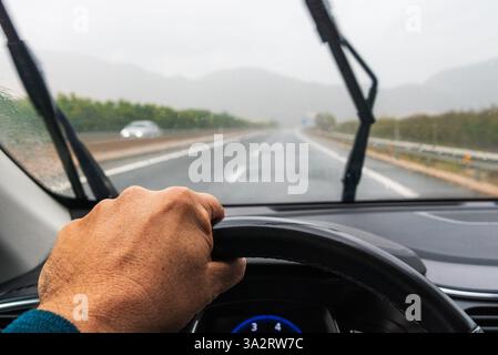 La main d'un homme saisissant fermement le volant d'une voiture un jour de pluie, conduisant sur une autoroute. Banque D'Images